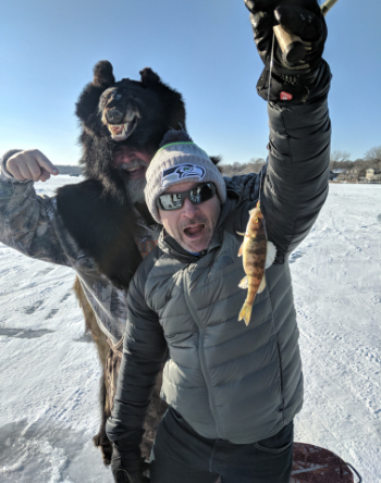 Man holding a small fish with another man standing behind him, wearing a bear skin as a hat